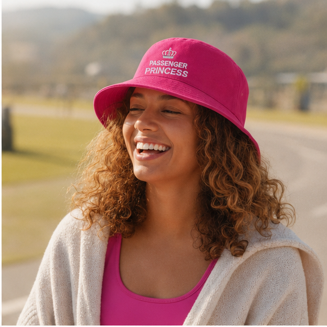 A woman outdoors wearing a bright pink bucket hat with white embroidered text reading 'PASSENGER PRINCESS' and a small crown above it. She is smiling, with a blurred road and greenery in the background.